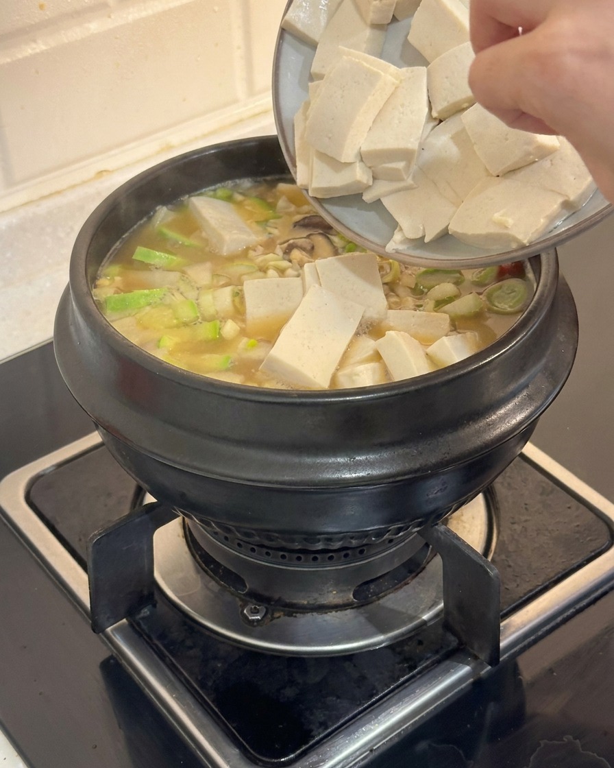 Mani che fanno scivolare cubetti di tofu bianco in una pentola fumante di stufato coreano