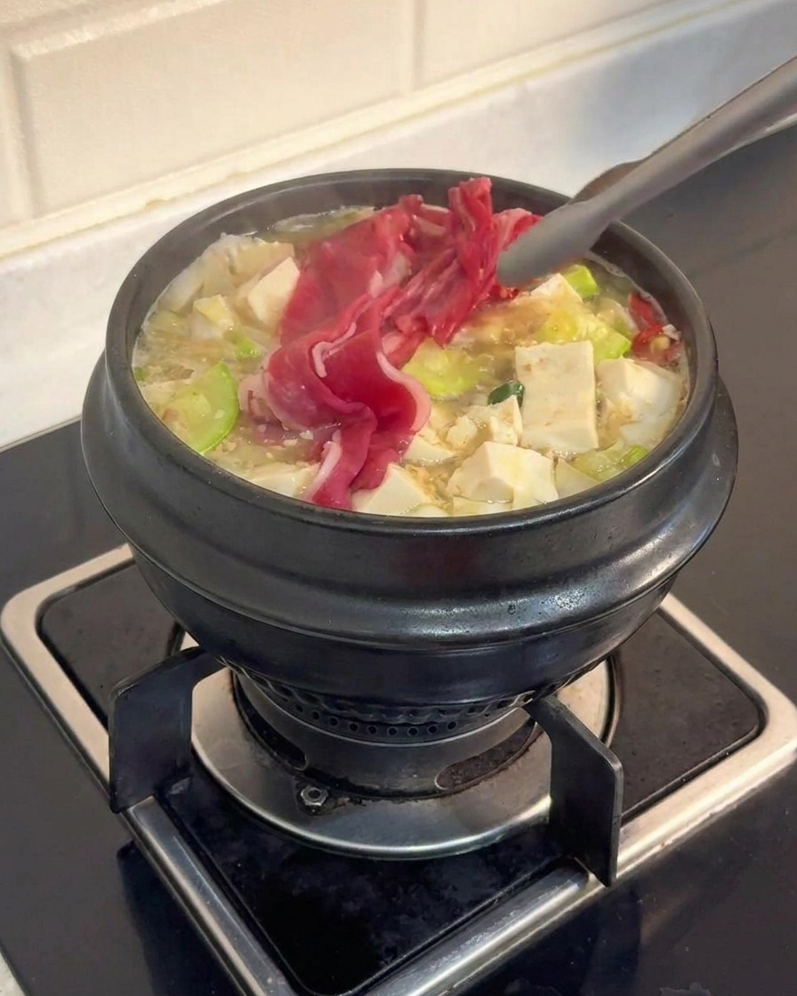 Pinze in metallo che immergono sottili fette di manzo marmorizzato in una zuppa di tofu e verdure