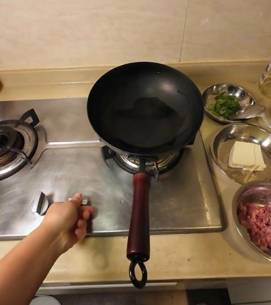 Una mano regola la manopola del fornello a gas sotto un wok nero contenente olio di semi trasparente.