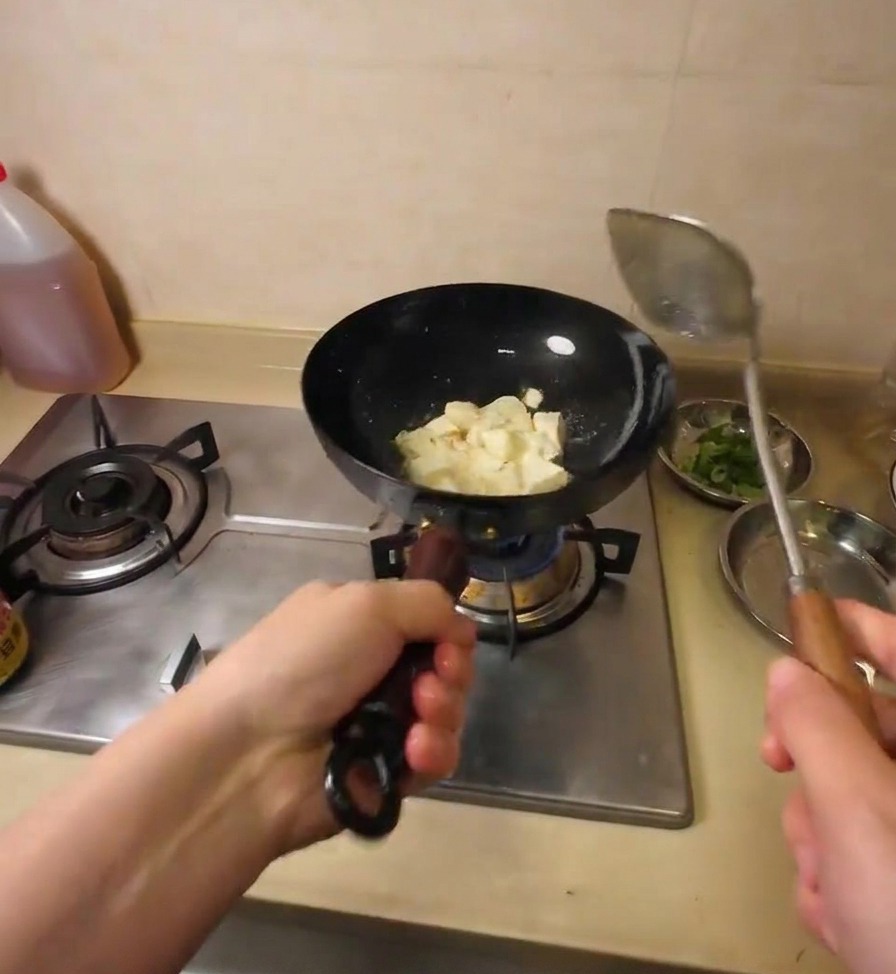 Una spatola di metallo rompe il blocco di tofu in cubetti irregolari mentre sfrigola nellolio caldo.