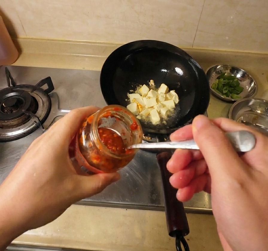 Un primo piano di una mano che aggiunge salsa al peperoncino rosso acceso in un wok con pezzi di tofu dorato.