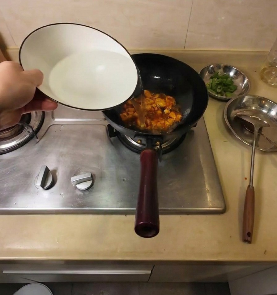 Acqua calda versata da una ciotola di ceramica bianca in un wok con tofu fritto e condimenti rossi.