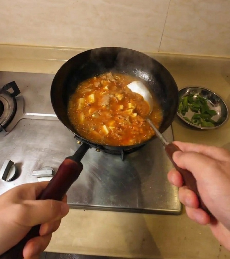 Una persona mescola una zuppa densa e arancione che bolle, contenente pezzi di manzo e tofu.
