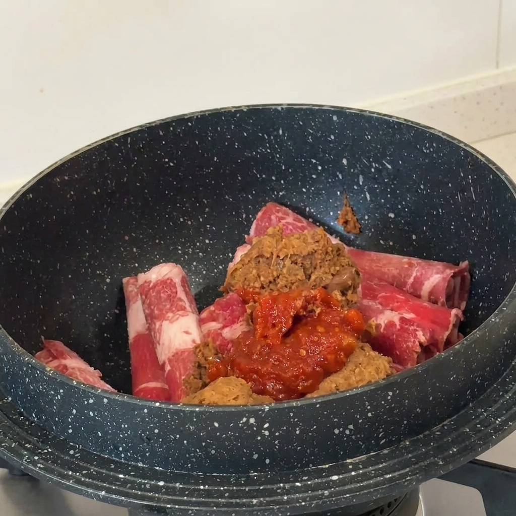 Pasta di soia marrone e pasta di peperoncino rosso sopra le fette di manzo crudo in una pentola scura.