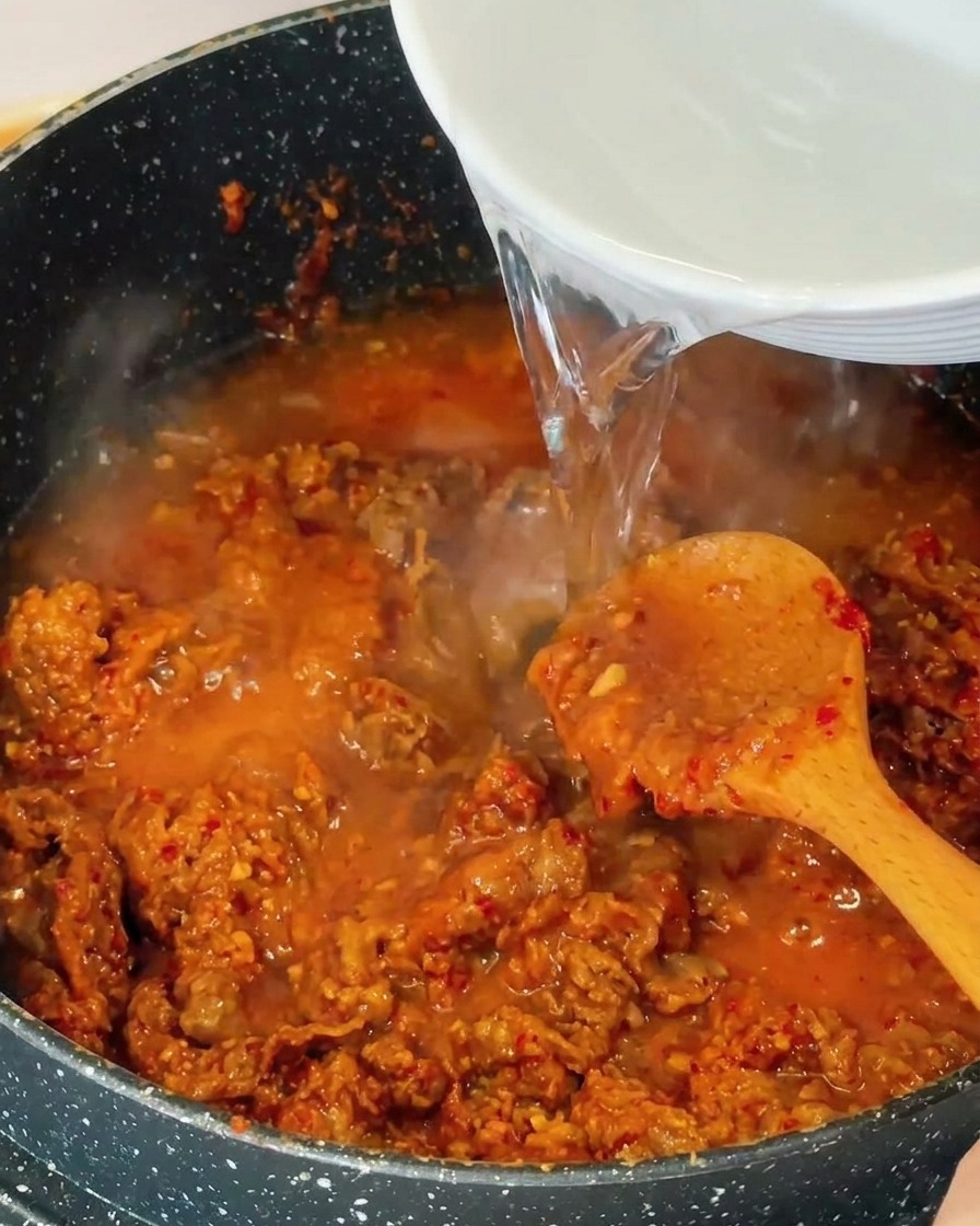 Acqua limpida versata da una ciotola bianca in una pentola con manzo rosolato e brodo rosso-marrone bollente.