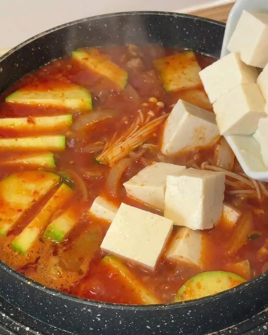 Cubetti di tofu bianco aggiunti a una pentola con zuppa bollente, zucchine e funghi enoki.
