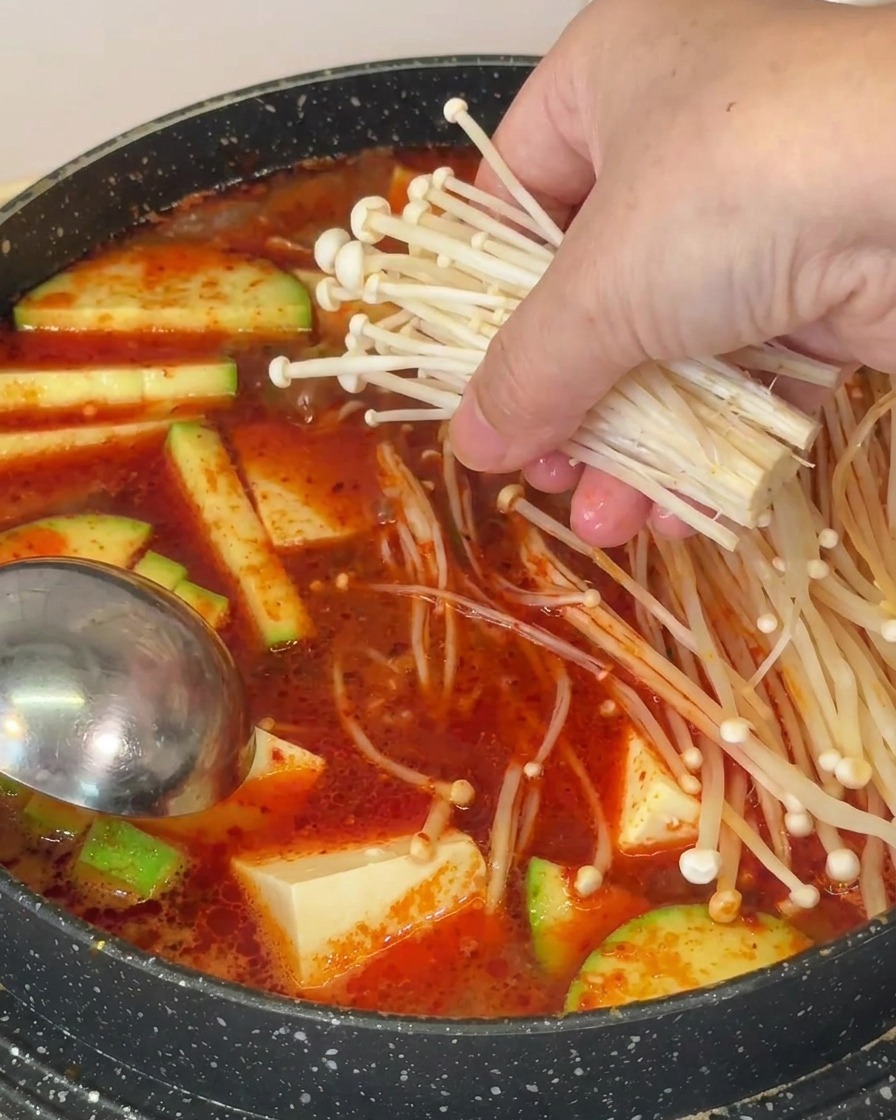 Una mano che mette un mazzetto di funghi enoki lunghi e sottili in una pentola di zuppa rossa con tofu e zucchine.