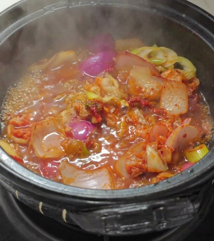 Acqua bollente versata in una pentola di pietra con kimchi e cipolle in un brodo rosso vibrante.