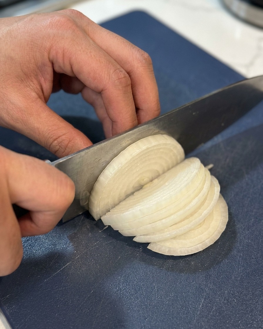 Un primo piano di mani che affettano una cipolla bianca a strisce sottili su un tagliere blu scuro.
