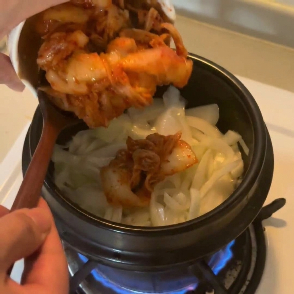 Kimchi di cavolo tritato versato da una ciotola di ceramica bianca nella pentola con il soffritto di cipolle.
