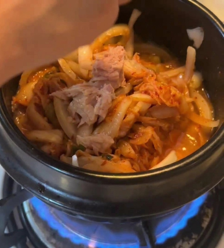 Pezzi di tonno in scatola aggiunti alla pentola con kimchi e cipolle sopra una fiamma a gas blu.