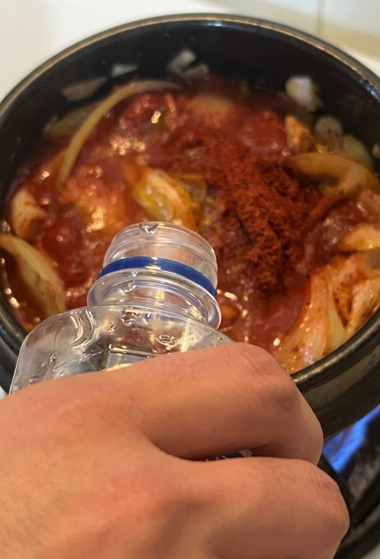 Acqua versata da una bottiglia trasparente nella pentola con lo stufato rosso vibrante sul fornello.