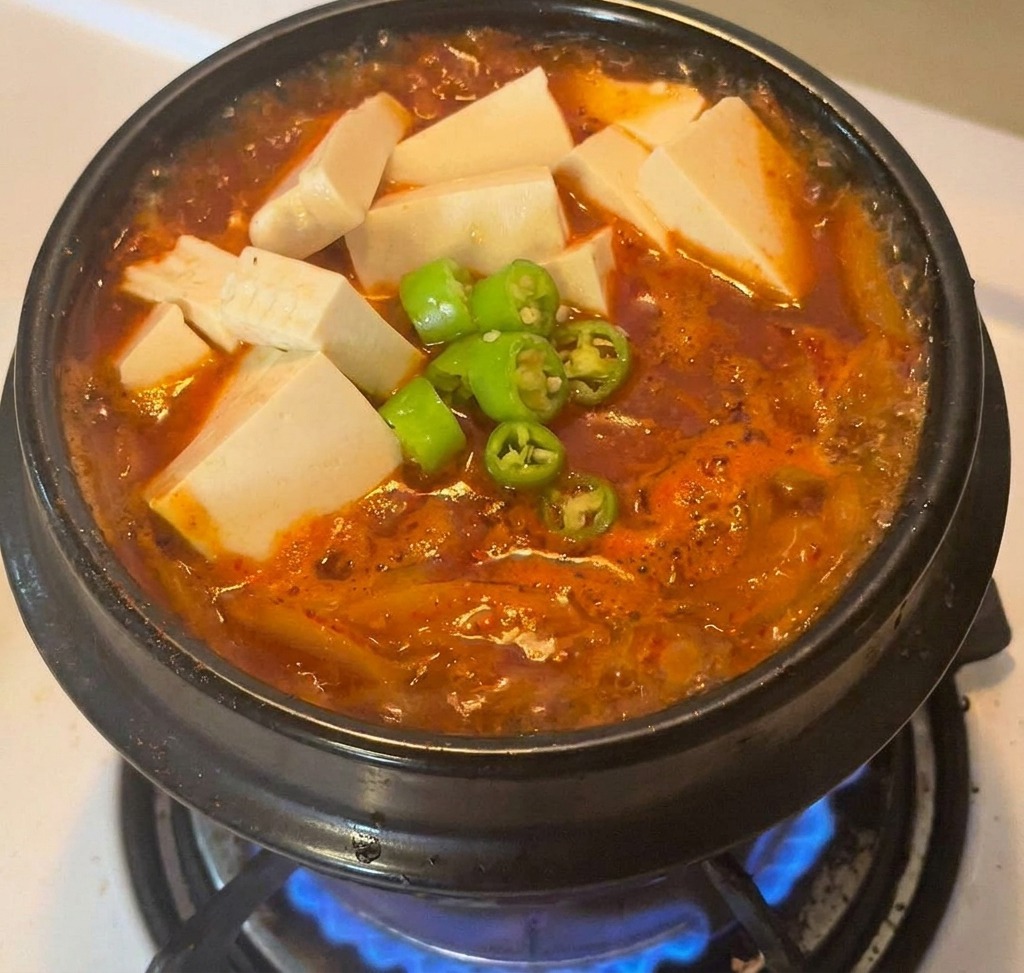 Una pentola di pietra con lo stufato rosso guarnito con cubetti di tofu e una manciata di peperoncini verdi affettati.