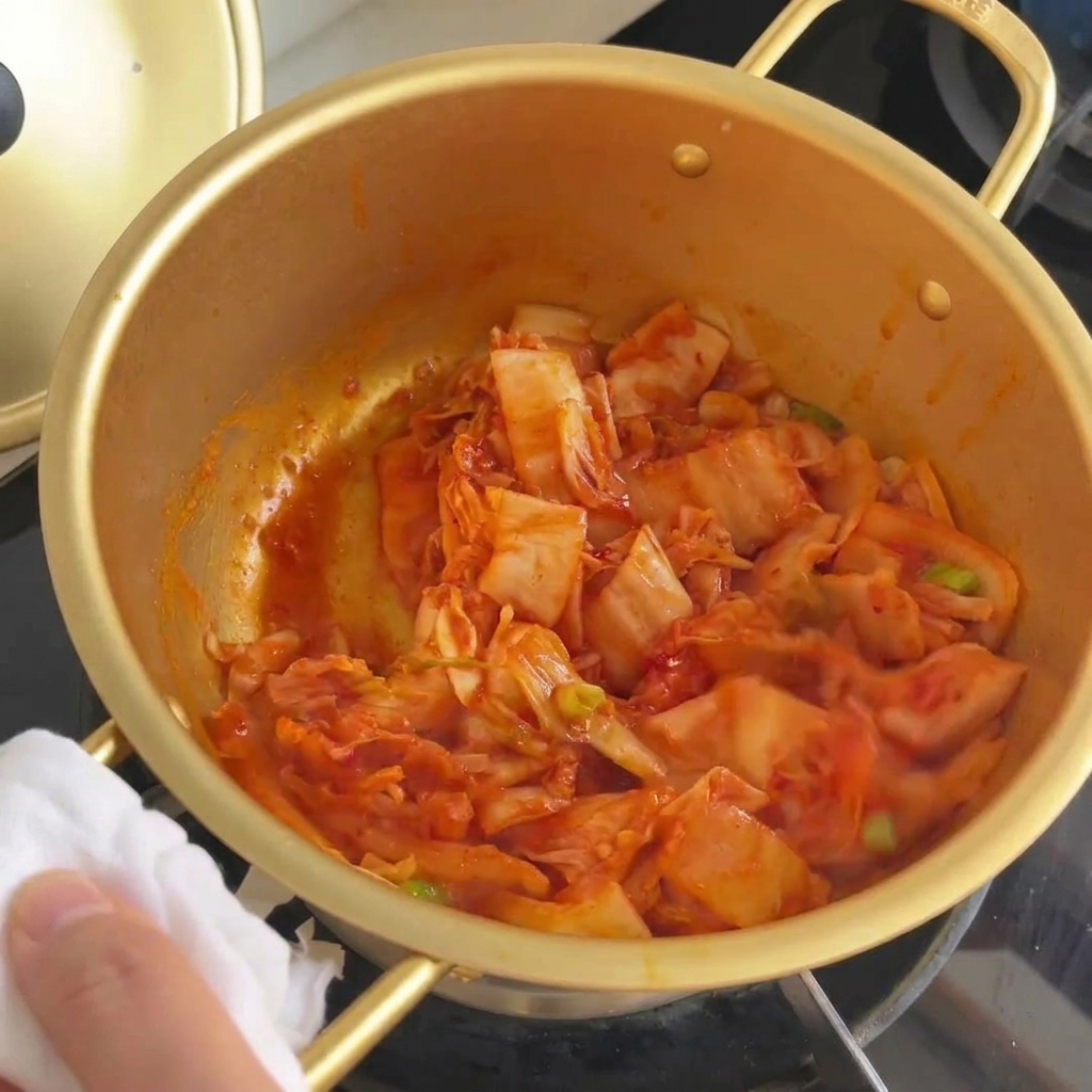 Kimchi e cipollotti saltati insieme in una pentola dorata finché il cavolo non appare traslucido.