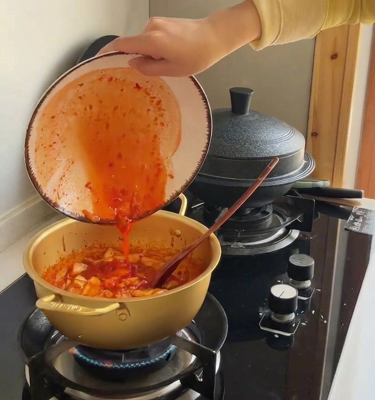 Succo di kimchi rosso vibrante versato da una ciotola in una pentola con cavolo saltato.