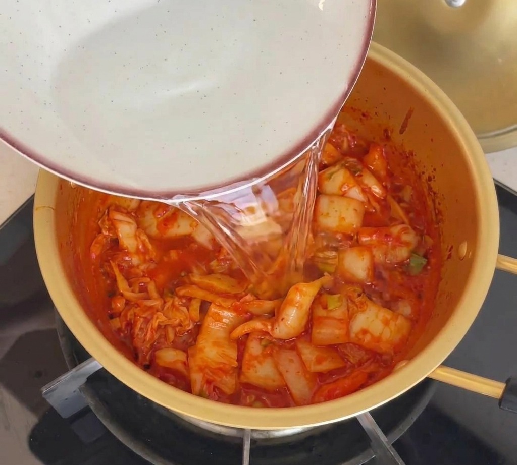 Acqua versata da una ciotola in una pentola dorata contenente il kimchi rosso saltato.