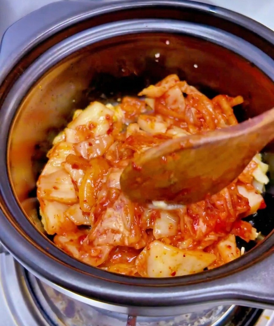 Il liquido di una scatoletta di tonno versato sopra un mix di kimchi tritato e aglio in una pentola di ceramica nera.