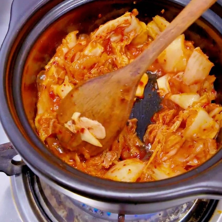 Una mano con un guanto nero aggiunge fette di cipolla bianca in una pentola contenente kimchi rosso.