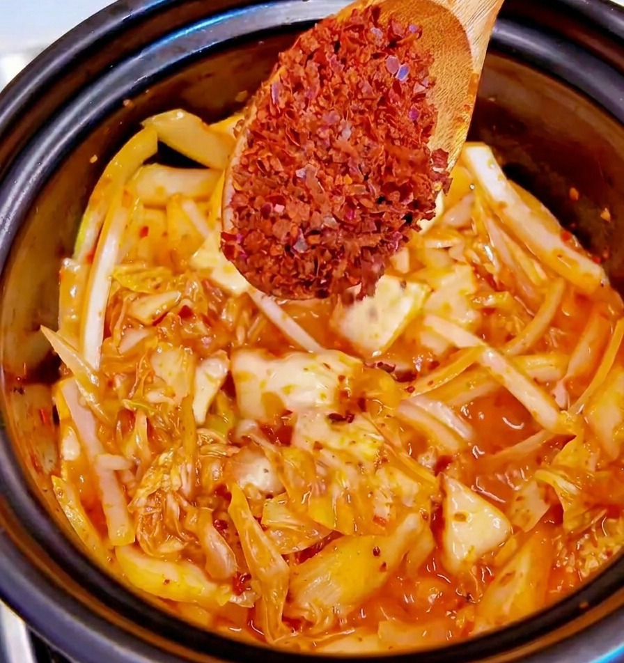 Un cucchiaio di legno con scaglie di peperoncino rosso sopra una pentola con kimchi e cipolle che soffriggono.