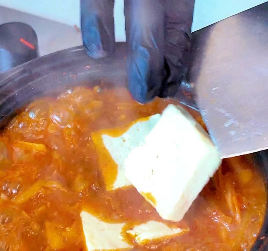 Fette di tofu bianco immerse in una zuppa di kimchi rossa e bollente usando una spatola.