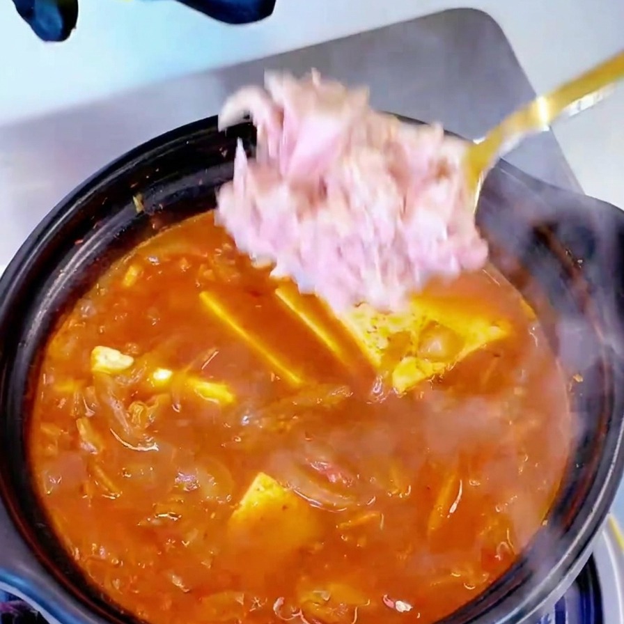 Pezzi di tonno in scatola aggiunti con un cucchiaio a una pentola di zuppa bollente di kimchi e tofu.