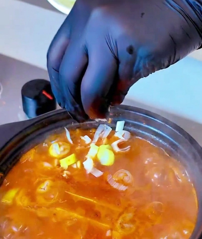 Una mano con un guanto nero sparge una manciata di cipollotti tritati in una pentola fumante di zuppa rosso-arancio.