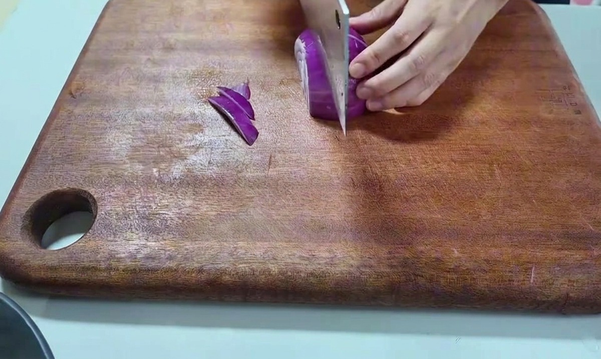 Una mano che tiene una cipolla rossa e la taglia con un coltello da cucina su un tagliere di legno.