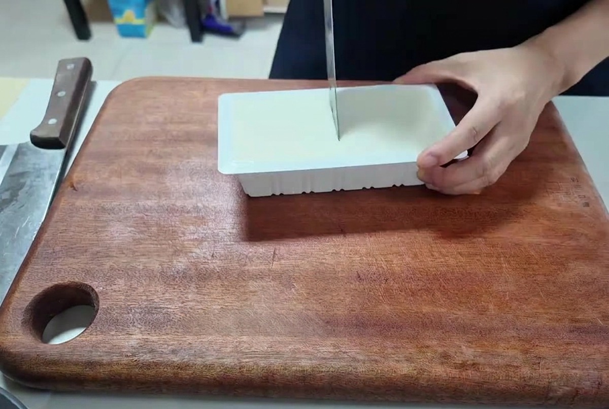 Una persona che usa un coltello per tagliare un panetto di tofu bianco a cubetti su un tagliere.