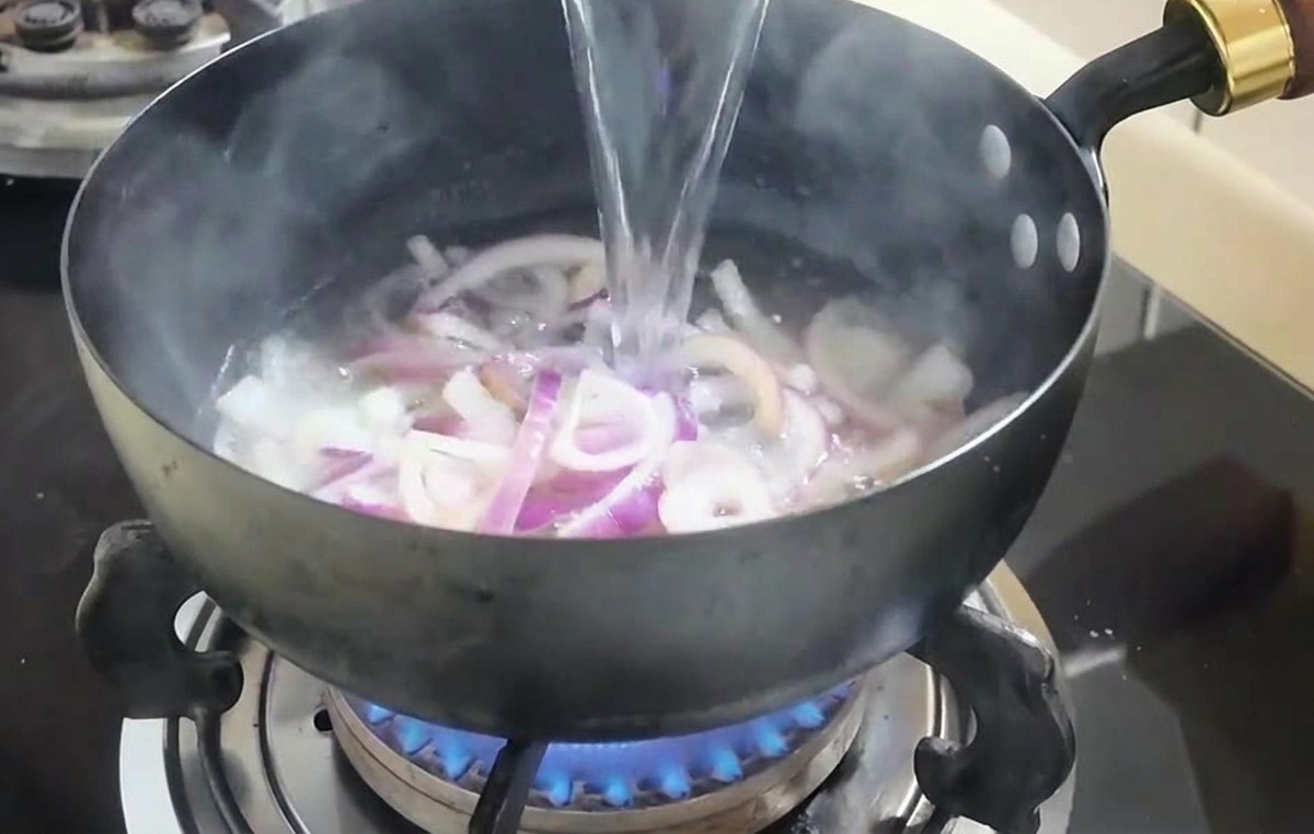 Un getto dacqua versato da una ciotola bianca in una padella con le cipolle soffritte.