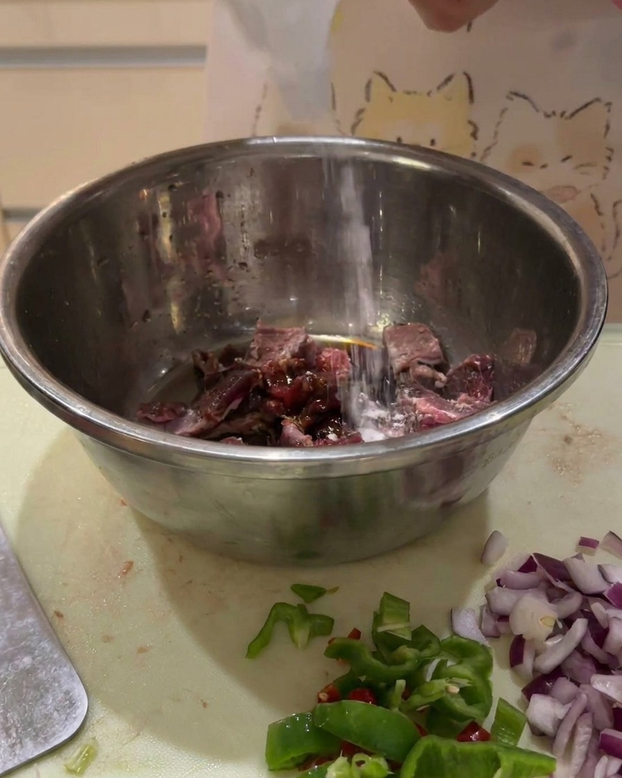 Salse scure versate da una bottiglia su cubetti di carne cruda in una ciotola di metallo.
