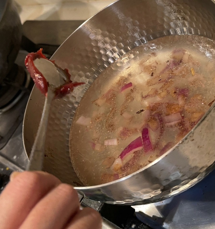 Un cucchiaio che immerge una dose generosa di pasta rossa densa nella pentola con acqua e cipolle.