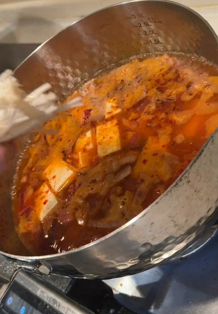 Una mano che aggiunge funghi enoki in una pentola di zuppa rossa ribollente con cubetti di tofu.