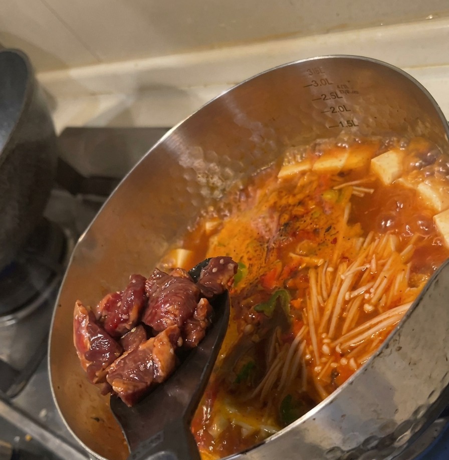 Cubetti di manzo crudo spinti da una spatola in una zuppa rossa bollente ricca di verdure.