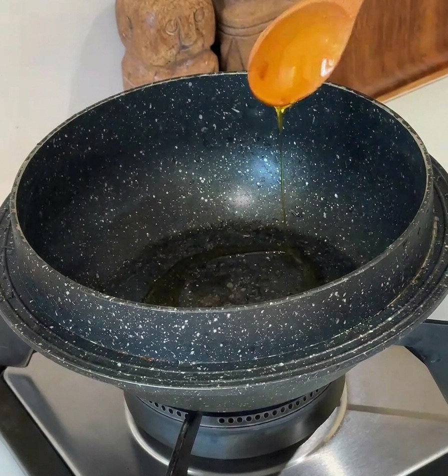 Olio di semi versato da un cucchiaio di legno chiaro in una pentola di pietra scura su un piano cottura moderno.