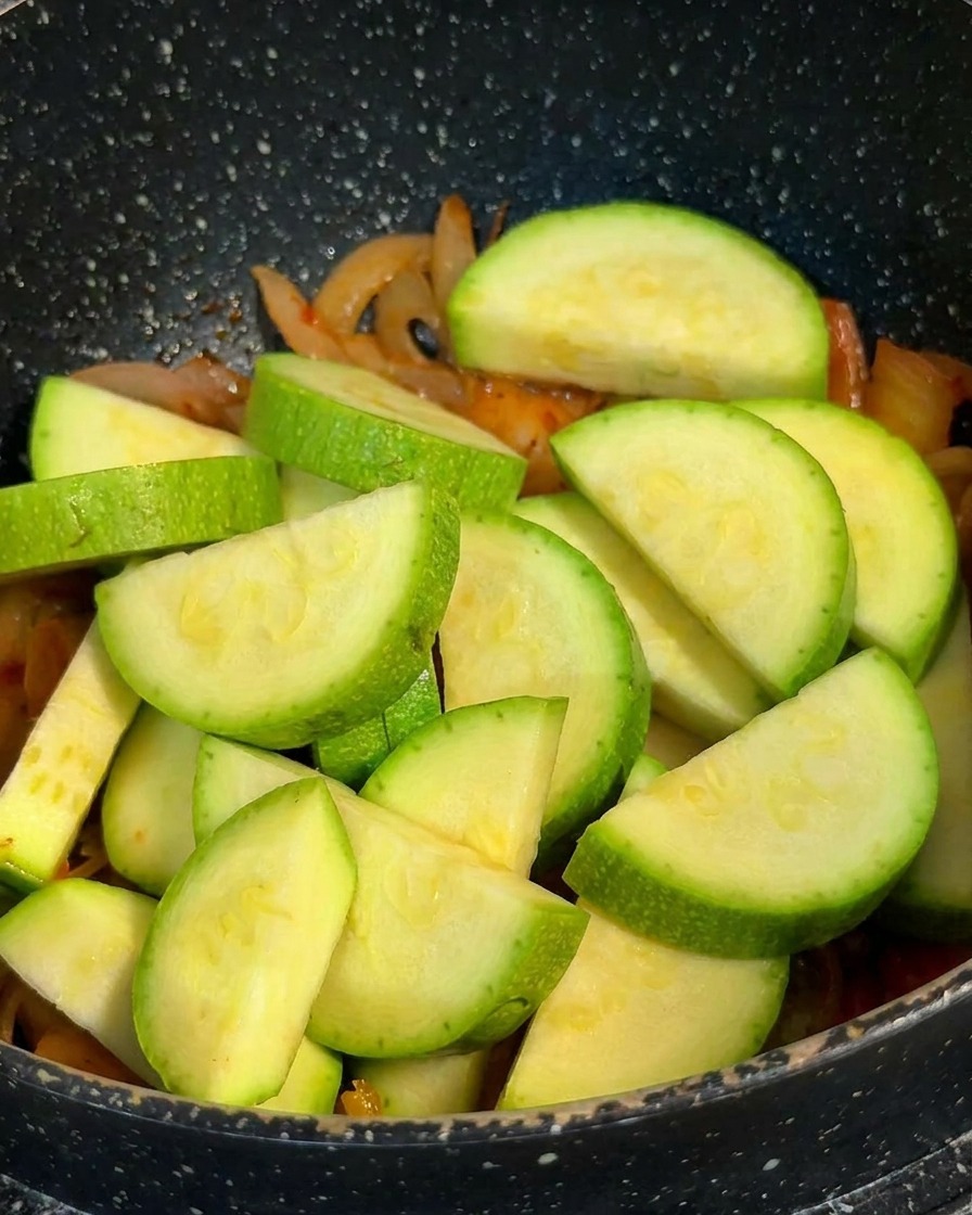 Fette di zucchina verde fresca a mezzaluna impilate in una pentola sopra una base di kimchi e cipolle.
