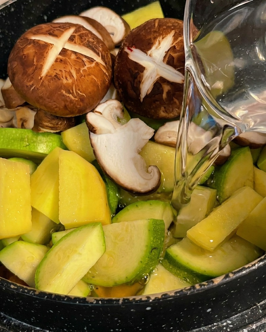 Acqua limpida versata da una caraffa su un mix colorato di zucchine, patate e funghi shiitake in una pentola.
