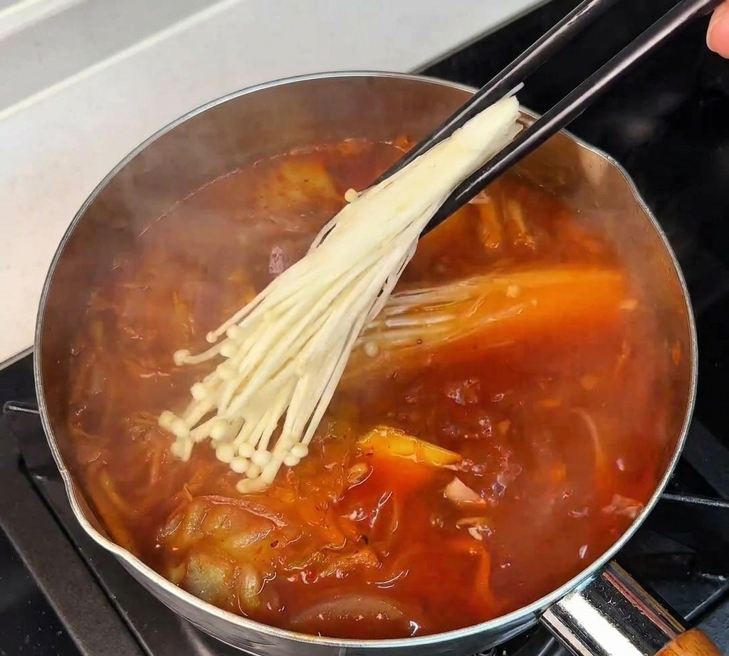 Una mano usa le bacchette per immergere un mazzetto di funghi enoki bianchi in una zuppa di kimchi rossa che bolle.