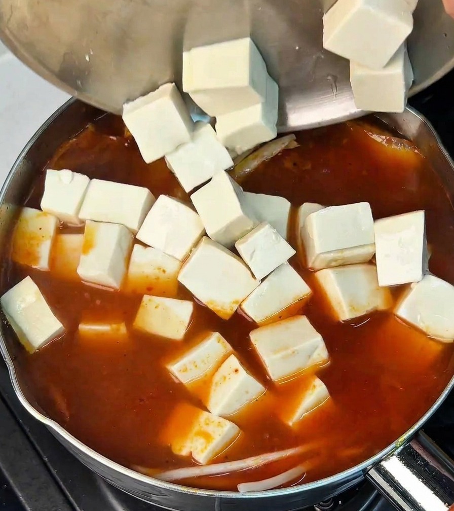 Cubetti di tofu bianco versati da una ciotola dacciaio in una pentola di brodo di kimchi rosso.