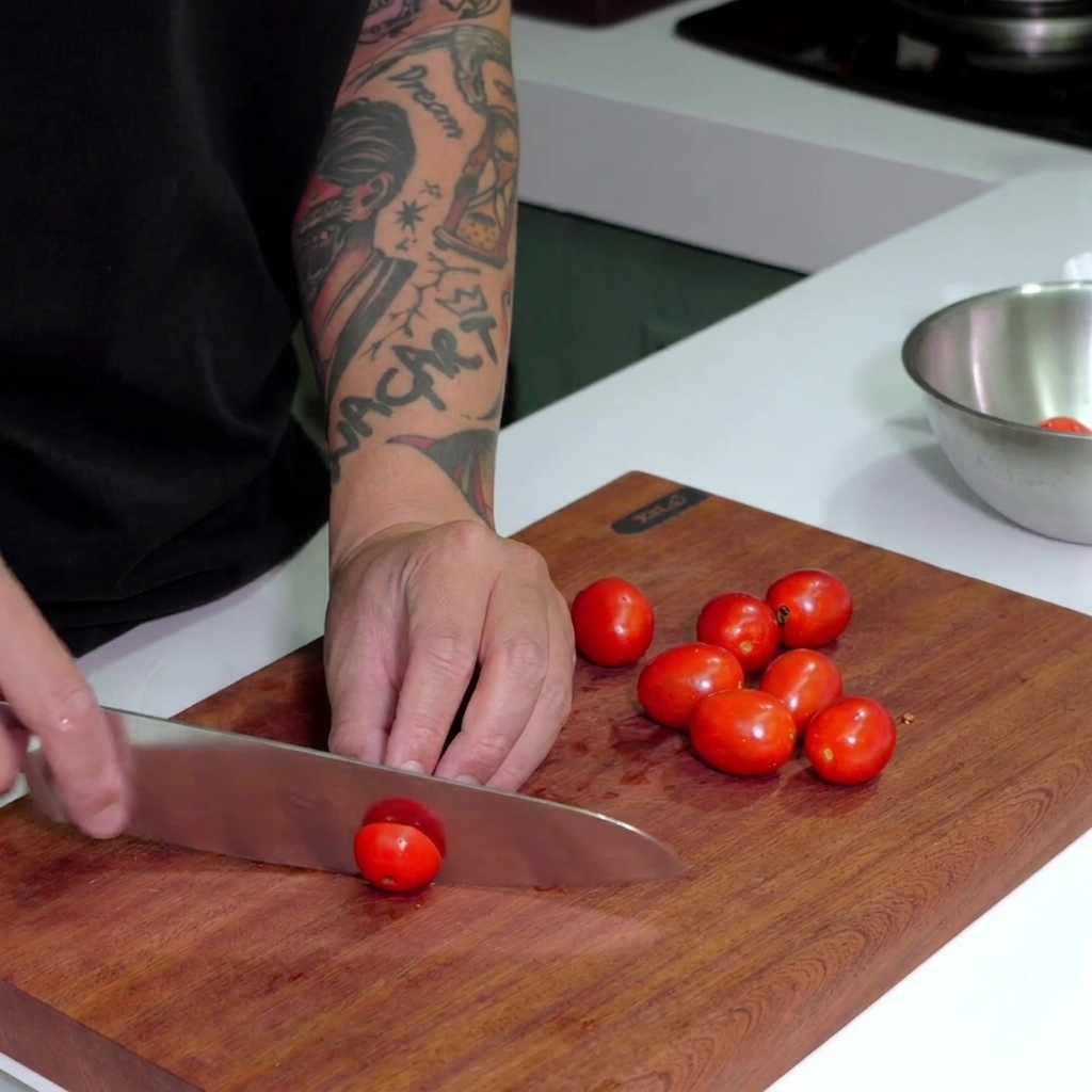 Un coltello da chef che taglia pomodorini rossi maturi su un tagliere di legno scuro.