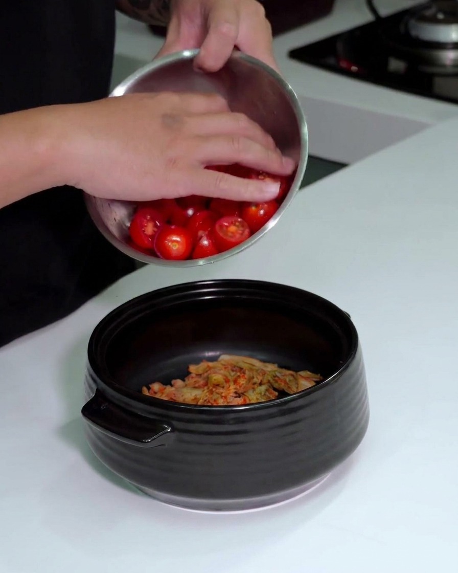 Una persona che versa una ciotola di pomodorini rossi affettati in una pentola nera che contiene già il kimchi.