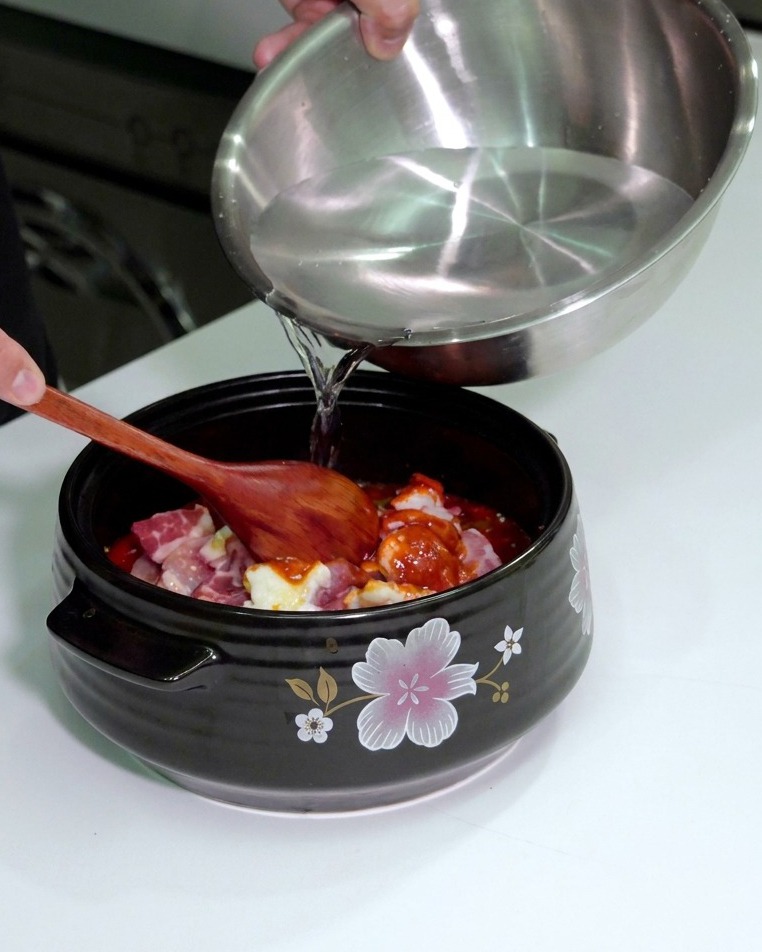 Persona che versa acqua da una ciotola dacciaio in una pentola di ceramica con carne e verdure condite.