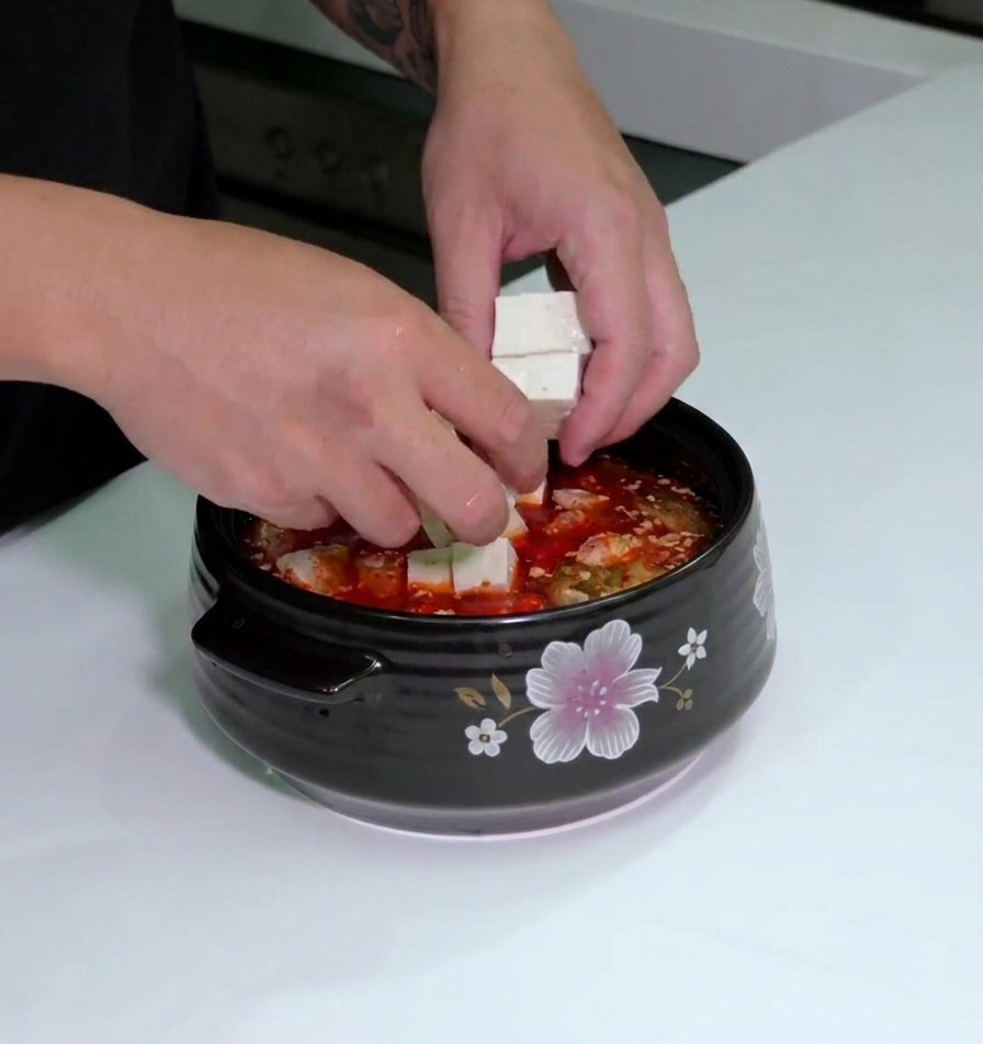Mani che posizionano cubetti di tofu bianco fresco in una pentola di ceramica con la base di zuppa rossa.