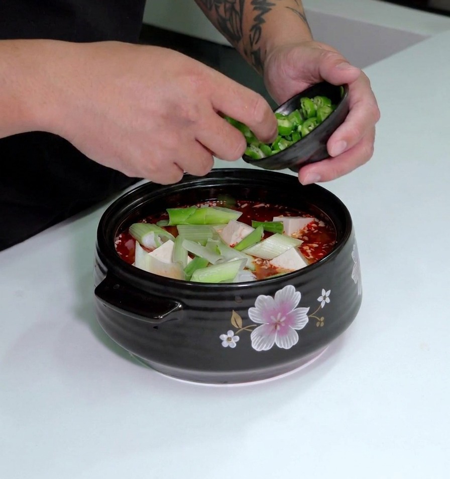 Peperoncini verdi affettati che vengono cosparsi sopra una pentola di zuppa di kimchi con tofu e porri.