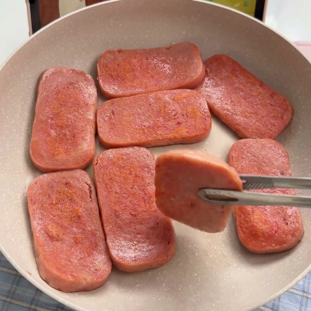 Sette fette rettangolari di carne in scatola rosa che rosolano in una padella antiaderente fino a diventare dorate.