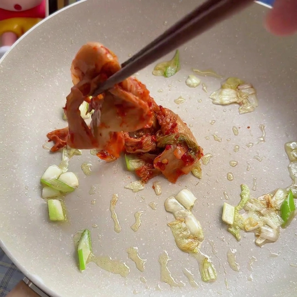 Delle bacchette che aggiungono kimchi di cavolo tritato in una padella insieme ai cipollotti già soffritti.