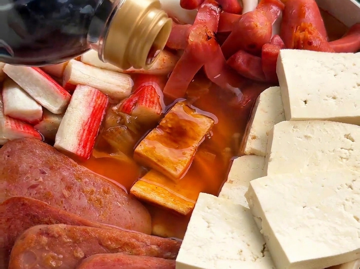 Salsa di soia scura versata nel brodo rosso che ribolle insieme a würstel, surimi e carne in scatola.