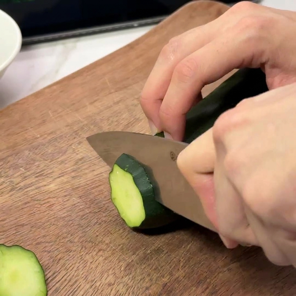 Primo piano di mani che usano un coltello da chef per affettare un cetriolo fresco a rondelle sottili su un tagliere di legno.