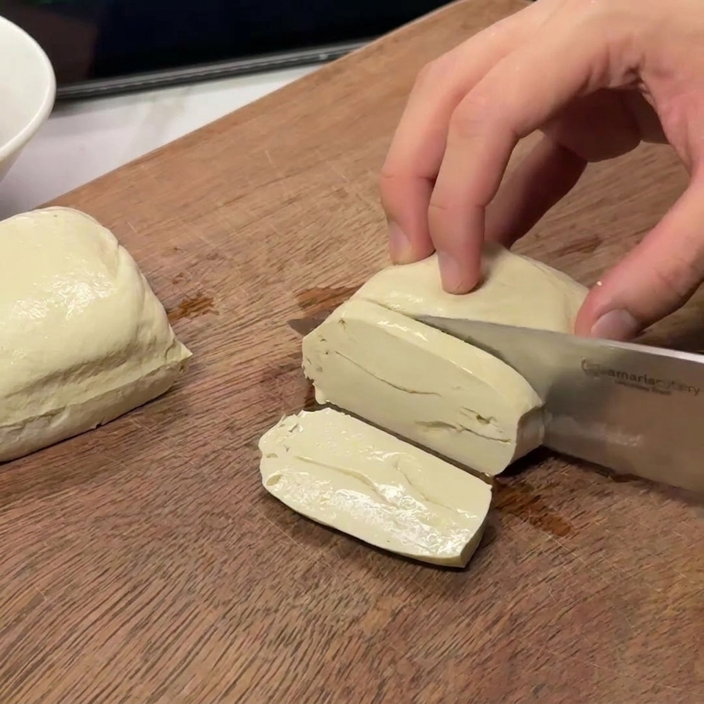 Mani che tagliano un blocco di tofu bianco sodo in fette rettangolari consistenti su una superficie di legno.