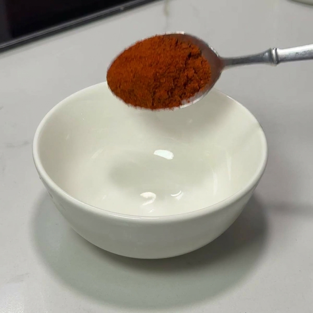 Un cucchiaio dargento che versa una manciata di peperoncino rosso brillante in polvere in una ciotola di ceramica bianca pulita.