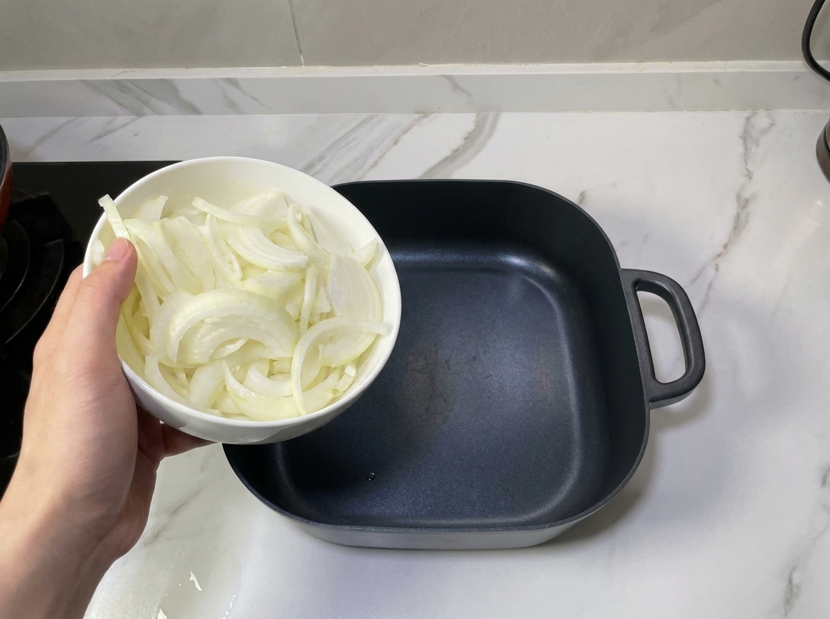 Una mano che tiene una ciotola di cipolle affettate sopra una pentola quadrata nera vuota, pronta per disporle sul fondo.